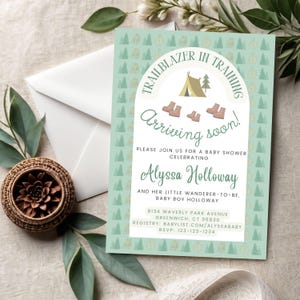 Puede incluir: Una invitación a un baby shower con un fondo verde menta y un patrón repetido de árboles y brújulas. El texto "Trailblazer in Training" se arquea sobre un gráfico de tienda de campaña y botas. La invitación dice "Arriving soon!" e incluye el nombre Alyssa Holloway.