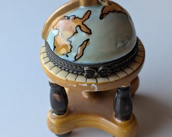 Puede incluir: Una pequeña caja esférica para baratijas con forma de globo terráqueo, con una superficie azul claro y continentes pintados en dorado. El globo descansa sobre un soporte beige de cuatro patas con detalles en marrón oscuro y una bisagra metálica.
