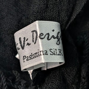 May include: Close-up of a label from a black Pashmina Silk scarf. The label is light gray with black text that reads "Vi Design" and "Pashmina Silk". The scarf has a textured appearance.