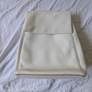 May include: Cream-colored leather backpack with a flap closure. The bag has a textured surface and a brown trim along the bottom edge. The backpack is sitting on a white, wrinkled fabric.