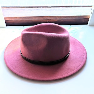 Puede incluir: Un sombrero fedora rosa polvoriento con ala plana y una banda marr&oacute;n oscuro. El sombrero tiene una forma cl&aacute;sica y estructurada y est&aacute; hecho de un material suave. Es un accesorio elegante.