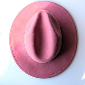 Puede incluir: Un sombrero fedora rosa polvoriento con ala ancha. El sombrero tiene una corona cl&aacute;sica en forma de l&aacute;grima y una cinta de grogr&eacute;n. El sombrero est&aacute; hecho de fieltro suave, un accesorio elegante.