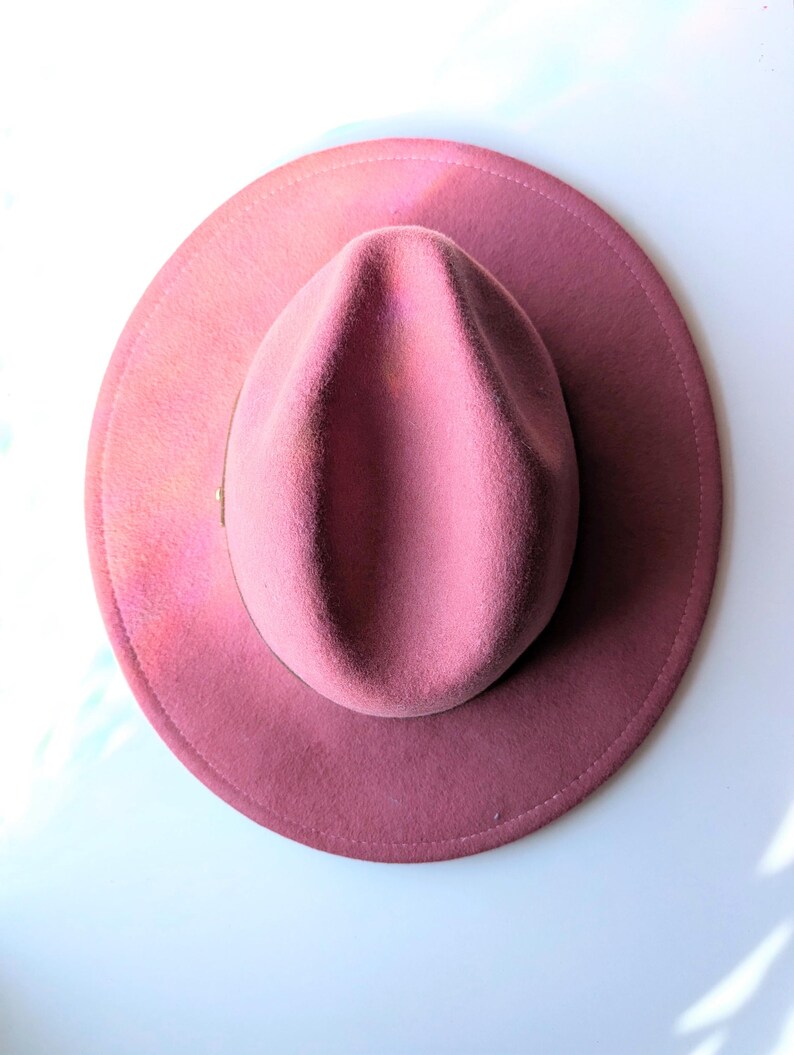 Puede incluir: Un sombrero fedora rosa polvoriento con ala ancha. El sombrero tiene una corona cl&aacute;sica en forma de l&aacute;grima y una cinta de grogr&eacute;n. El sombrero est&aacute; hecho de fieltro suave, un accesorio elegante.