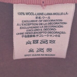 Puede incluir: Primer plano de una etiqueta de tela rosa con texto en varios idiomas, incluidos ingl&eacute;s, franc&eacute;s y espa&ntilde;ol. La etiqueta dice "100% LANA" e incluye s&iacute;mbolos de cuidado y las palabras "SPOT CLEAN AS NEEDED."