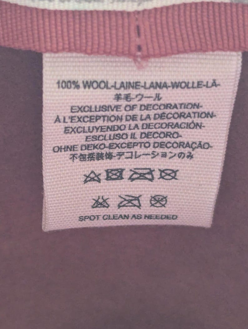 Puede incluir: Primer plano de una etiqueta de tela rosa con texto en varios idiomas, incluidos ingl&eacute;s, franc&eacute;s y espa&ntilde;ol. La etiqueta dice "100% LANA" e incluye s&iacute;mbolos de cuidado y las palabras "SPOT CLEAN AS NEEDED."