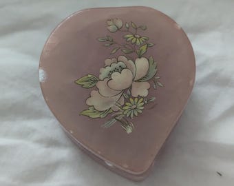 May include: A heart-shaped, pink trinket box with a floral design. The box features a large, cream-colored flower with green leaves and smaller yellow flowers. The box is made of a smooth material and has a slightly glossy finish.