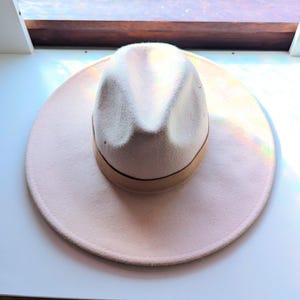 Może przedstawiać: Jasnor&oacute;żowy kapelusz typu fedora z kremową koroną i brązową opaską. Kapelusz ma szerokie rondo i klasyczny, stylowy design. Ten kapelusz jest idealny, aby dodać odrobinę elegancji do każdego stroju.