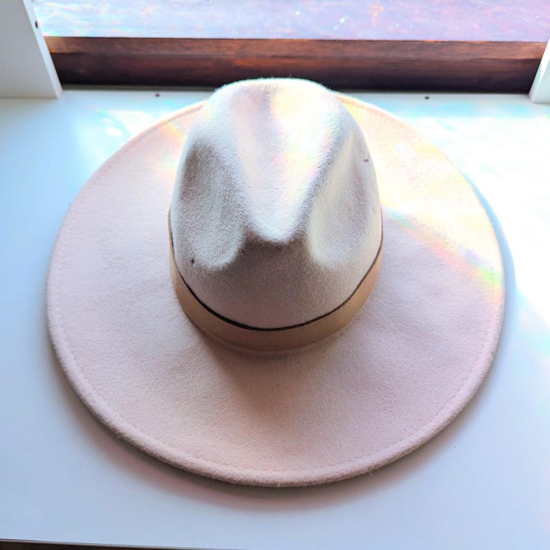 Może przedstawiać: Jasnor&oacute;żowy kapelusz typu fedora z kremową koroną i brązową opaską. Kapelusz ma szerokie rondo i klasyczny, stylowy design. Ten kapelusz jest idealny, aby dodać odrobinę elegancji do każdego stroju.