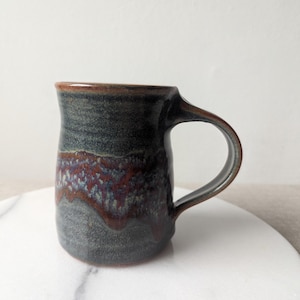 May include: A handmade ceramic mug with a dark blue and brown glaze. The mug features a rounded body, a sturdy handle, and a band of reddish-brown and purple hues around the middle. The mug is displayed on a white surface.