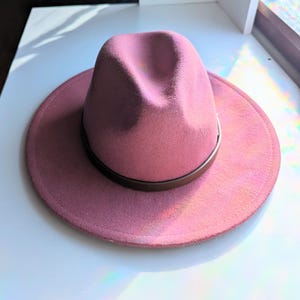 Puede incluir: Un sombrero fedora rosa polvoriento con una banda de cuero marr&oacute;n. El sombrero tiene un ala ancha y una forma de corona cl&aacute;sica. Este elegante sombrero es perfecto para a&ntilde;adir un toque de sofisticaci&oacute;n a cualquier atuendo.