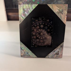 May include: A small, rectangular picture frame with a black center and iridescent, shell-like accents. The frame has a geometric design with a black diamond shape in the center. The iridescent sections have a mix of colors, including green, blue, and silver.