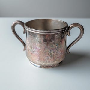 May include: A small, two-handled silver cup with a rounded body and a wide rim. The metal surface shows significant signs of oxidation, featuring a mottled patina with visible patches of iridescent pink, blue, and dark gray tarnish throughout.