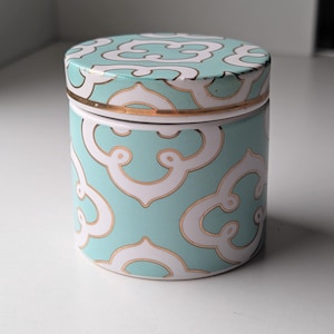 May include: A cylindrical ceramic jar with a removable lid, featuring a repeating quatrefoil pattern in white and gold outlines against a light turquoise background. The container has a smooth finish and a gold-toned rim around the lid edge.