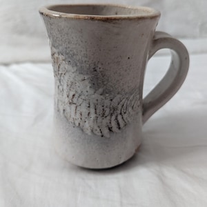May include: A handmade ceramic mug with a textured, off-white glaze. The mug features a dark brown, feather-like design on the front and a curved handle. The rim and handle have a darker, speckled finish. The mug is approximately 4 inches tall.