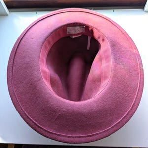 Puede incluir: Un sombrero fedora burdeos con ala ancha. El interior del sombrero es visible, mostrando un forro rosa-rojizo y una etiqueta. El sombrero est&aacute; hecho de un material similar al fieltro y tiene un dise&ntilde;o cl&aacute;sico y atemporal.
