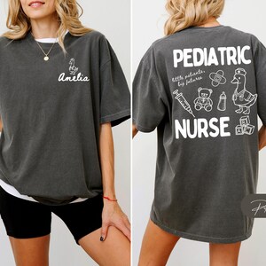 Puede incluir: Camiseta gris oscuro con texto y gr&aacute;ficos blancos. La parte delantera presenta un dibujo de pato y el nombre "Amelia". La parte trasera muestra "PEDIATRIC NURSE" con ilustraciones m&eacute;dicas y para beb&eacute;s. La camiseta tiene un ajuste holgado y c&oacute;modo.