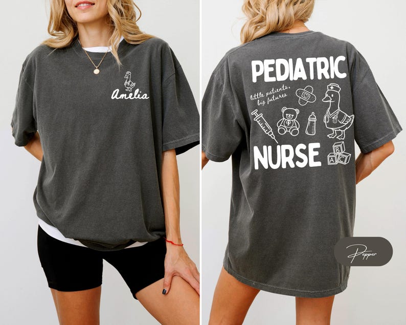 Puede incluir: Camiseta gris oscuro con texto y gr&aacute;ficos blancos. La parte delantera presenta un dibujo de pato y el nombre "Amelia". La parte trasera muestra "PEDIATRIC NURSE" con ilustraciones m&eacute;dicas y para beb&eacute;s. La camiseta tiene un ajuste holgado y c&oacute;modo.