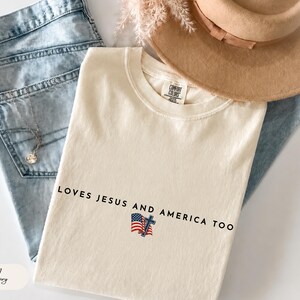 Pu&ograve; includere: T-shirt color avorio con la scritta "LOVES JESUS AND AMERICA TOO" e una piccola bandiera americana e grafica a croce. La maglietta &egrave; piegata sopra jeans blu, con un cappello e gioielli visibili sullo sfondo.