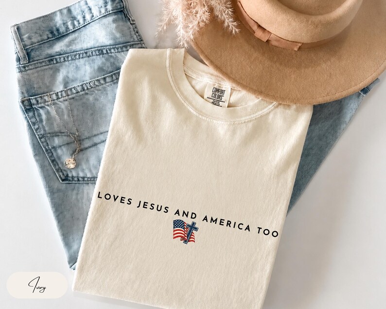 Pu&ograve; includere: T-shirt color avorio con la scritta "LOVES JESUS AND AMERICA TOO" e una piccola bandiera americana e grafica a croce. La maglietta &egrave; piegata sopra jeans blu, con un cappello e gioielli visibili sullo sfondo.