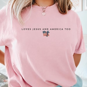 Pu&ograve; includere: T-shirt rosa chiaro con la scritta nera "LOVES JESUS AND AMERICA TOO". Sotto il testo c'&egrave; una piccola grafica della bandiera americana e una croce. La maglietta &egrave; a tinta unita e ha maniche corte.