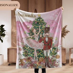 May include: A personalized pink and white blanket featuring a Christmas scene with a ballerina, a nutcracker, and a decorated Christmas tree. The blanket has the name "Amanda" in gold script and the word "PERSONALIZE" in a tan oval.