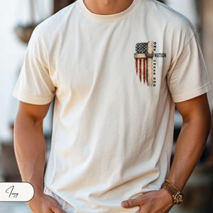 May include: An ivory short-sleeve t-shirt featuring a graphic on the upper left chest. The design depicts a wooden cross overlaid with a distressed American flag pattern and the text "ONE NATION UNDER GOD" printed in black capital letters.