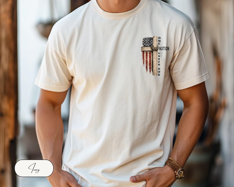 May include: An ivory short-sleeve t-shirt featuring a graphic on the upper left chest. The design depicts a wooden cross overlaid with a distressed American flag pattern and the text "ONE NATION UNDER GOD" printed in black capital letters.
