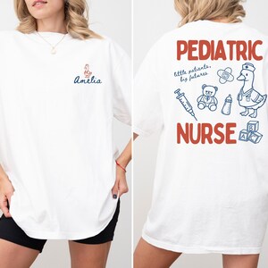 Puede incluir: Camiseta blanca con "Amelia" y un gr&aacute;fico de pato en la parte delantera. La parte trasera presenta "Pediatric Nurse" en texto rojo, junto con ilustraciones de un pato, un oso de peluche y suministros m&eacute;dicos en azul.