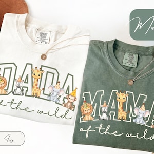 May include: Two t-shirts, one ivory and one green, with the text "DADA of the wild" and "MAMA of the wild" in white. Each shirt features cartoon animals wearing party hats. A gold necklace is draped on each shirt.