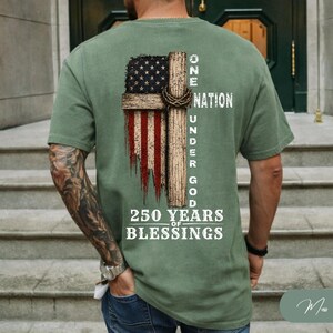 May include: An olive green short-sleeve t-shirt featuring a graphic on the back of a wooden cross overlaid with a distressed American flag. The text reads ONE NATION UNDER GOD 250 YEARS OF BLESSINGS in white, stylized, block-style lettering.