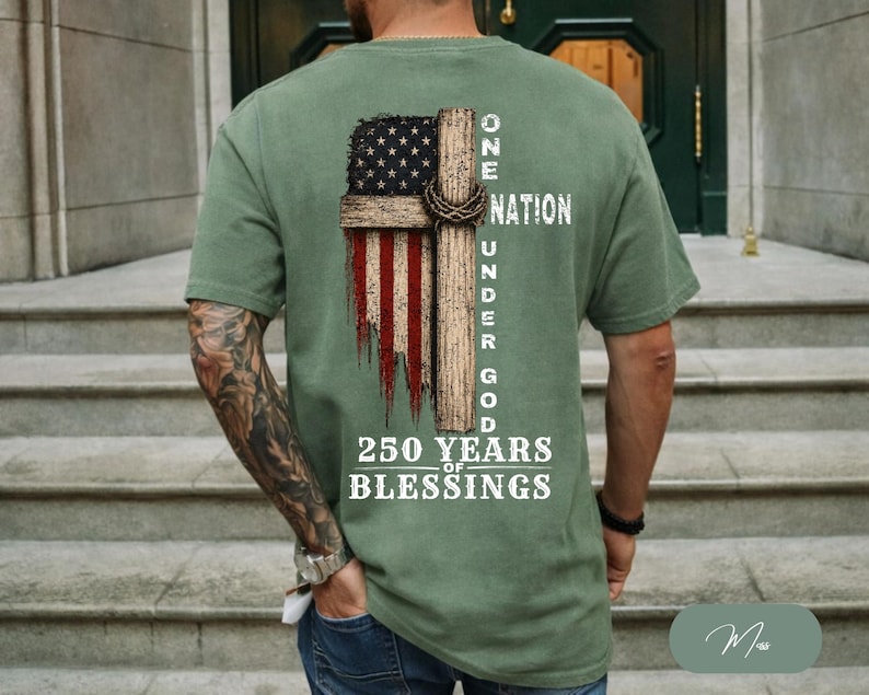 May include: An olive green short-sleeve t-shirt featuring a graphic on the back of a wooden cross overlaid with a distressed American flag. The text reads ONE NATION UNDER GOD 250 YEARS OF BLESSINGS in white, stylized, block-style lettering.