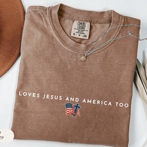 Pu&ograve; includere: Una maglietta marrone Comfort Colors con la scritta bianca "LOVES JESUS AND AMERICA TOO". La maglietta presenta una piccola grafica della bandiera americana e una croce. Una collana d'argento con un ciondolo &egrave; drappeggiata sulla maglietta.
