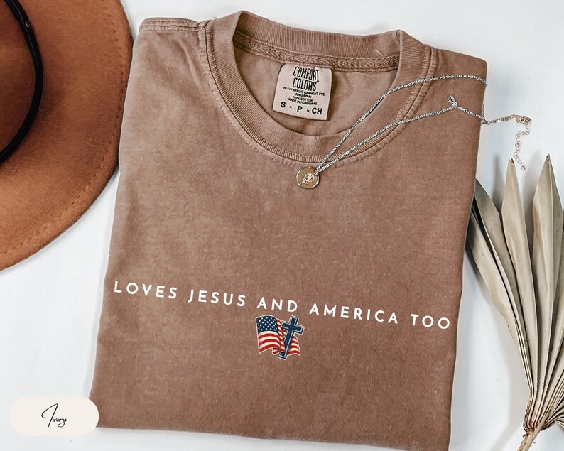 Pu&ograve; includere: Una maglietta marrone Comfort Colors con la scritta bianca "LOVES JESUS AND AMERICA TOO". La maglietta presenta una piccola grafica della bandiera americana e una croce. Una collana d'argento con un ciondolo &egrave; drappeggiata sulla maglietta.