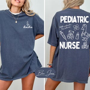 Puede incluir: Camiseta azul oscuro con la frase "Pediatric Nurse" e ilustraciones m&eacute;dicas, adem&aacute;s de un pato con gorro de enfermera. La parte delantera muestra el nombre "Amelia" y un peque&ntilde;o dibujo de pato.
