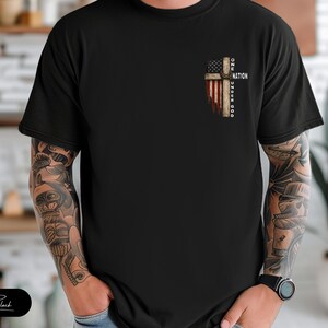 May include: A black short-sleeved t-shirt featuring a graphic on the left chest. The design depicts a wooden cross overlaid with an American flag pattern and the text ONE NATION UNDER GOD written vertically along the side of the cross.