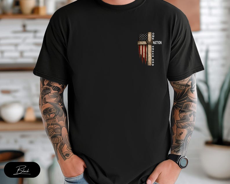 May include: A black short-sleeved t-shirt featuring a graphic on the left chest. The design depicts a wooden cross overlaid with an American flag pattern and the text ONE NATION UNDER GOD written vertically along the side of the cross.