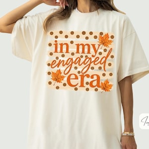 May include: An ivory short-sleeve t-shirt featuring a graphic print with brown polka dots, orange maple leaves, and the text in my engaged era in orange cursive and block lettering centered on the front of the garment.