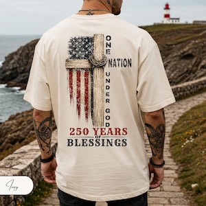 May include: An ivory short-sleeve t-shirt featuring a graphic on the back. The design includes a distressed American flag, a wooden cross with a crown of thorns, and the text: ONE NATION UNDER GOD 250 YEARS OF BLESSINGS in black and red.