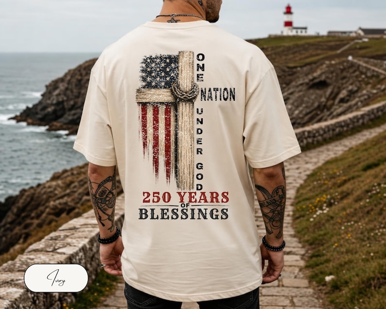 May include: An ivory short-sleeve t-shirt featuring a graphic on the back. The design includes a distressed American flag, a wooden cross with a crown of thorns, and the text: ONE NATION UNDER GOD 250 YEARS OF BLESSINGS in black and red.