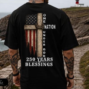May include: A black t-shirt featuring a graphic on the back. The design displays a wooden cross overlaid with a distressed American flag, a crown of thorns, and the text ONE NATION UNDER GOD 250 YEARS OF BLESSINGS in white capital letters.