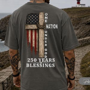 May include: A charcoal gray t-shirt featuring a back graphic of a distressed American flag integrated with a wooden cross and a crown of thorns. The text reads ONE NATION UNDER GOD and 250 YEARS OF BLESSINGS in a white, weathered, serif font.