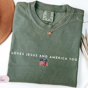 Pu&ograve; includere: T-shirt verde oliva con la scritta bianca "LOVES JESUS AND AMERICA TOO". Il design include una bandiera americana e una croce. Una collana d'oro con un ciondolo &egrave; appoggiata sulla maglietta.