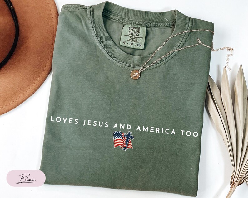 Pu&ograve; includere: T-shirt verde oliva con la scritta bianca "LOVES JESUS AND AMERICA TOO". Il design include una bandiera americana e una croce. Una collana d'oro con un ciondolo &egrave; appoggiata sulla maglietta.
