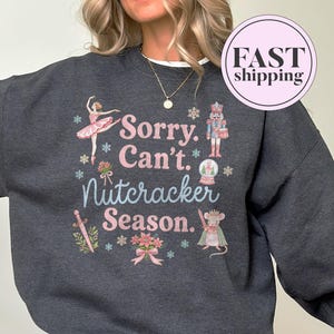 May include: A dark heather gray crewneck sweatshirt featuring a graphic print with the text Sorry. Can't. Nutcracker Season. The design includes illustrations of a ballerina, a nutcracker, a snow globe, a sword, a mouse, and poinsettia flowers.