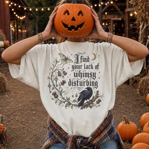 Könnte beinhalten: Ein cremefarbenes Kurzarm-T-Shirt mit einem Grafikdruck aus einer schwarzen Krähe, einer Motte, einer Mondsichel und Blumenranken. Der Text lautet, I find your lack of whimsy disturbing. Ein kariertes Flanellhemd ist um die Taille gebunden.
