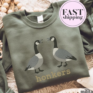 Könnte beinhalten: Militärgrünes Sweatshirt mit gestickten Gänsen, die sich gegenüberstehen. Unter den Gänsen ist das Wort "honkers" in Gold gestickt. Das Sweatshirt hat einen Rundhalsausschnitt.