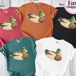 May include: Five short-sleeved t-shirts in crimson, yam, black, blue spruce, and white. Each shirt has a detailed duck illustration. The shirts are displayed on a light wood-grain background. A pink circle with "Fast Shipping" is in the upper right.