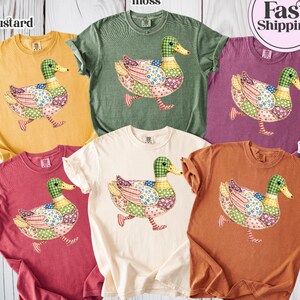 May include: A selection of Comfort Colors t-shirts in colours such as mustard, moss, berry, crimson, ivory, and yam. Each short-sleeved shirt displays a patchwork duck design. The image also includes a "Fast Shipping" graphic.