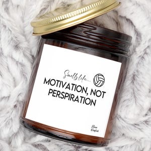 May include: A brown glass jar candle with a gold lid, featuring a white label with the text "Smells like... MOTIVATION, NOT PERSPIRATION" and a volleyball graphic. The candle sits on a fluffy, white and gray surface.