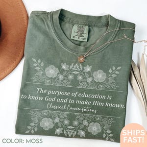 Pode incluir: Camiseta verde musgo com o texto "The purpose of education is to know God and to make Him known. Classical Conversations." O design floral envolve o texto. Um colar de ouro está na camisa.